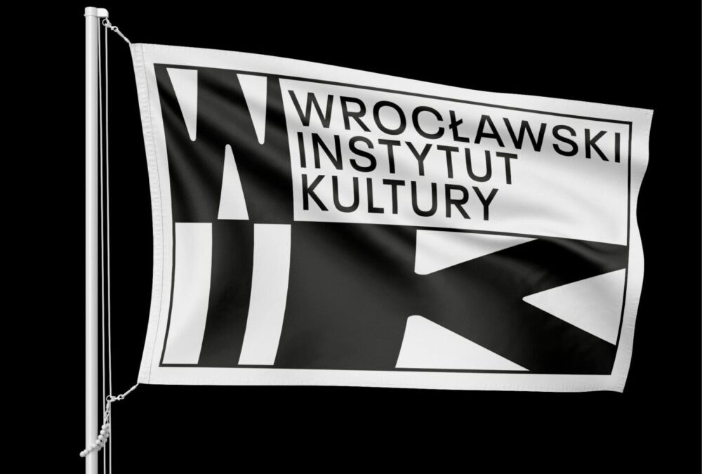 Grafika, czarno-biały logotyp Wrocławskiego Instytutu Kultury, osadzona na fladze. Logotyp składa się z czarnych liter WIK oraz napisu Wrocławski Instytut Kultury